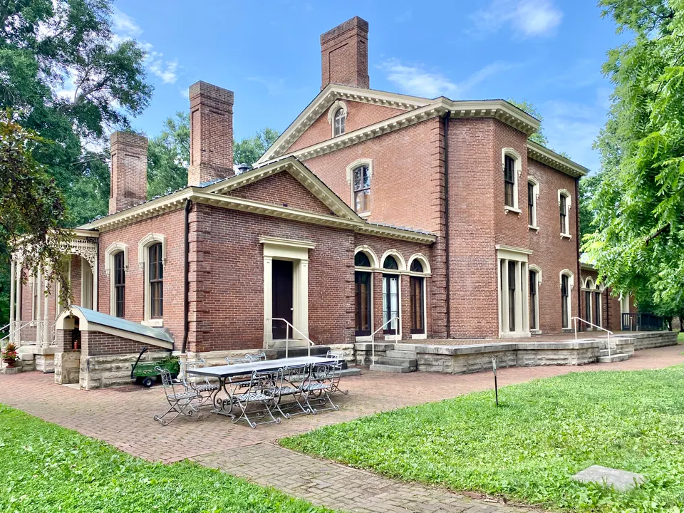 The rear of the red brick Ashland mansion in July 2023, with its stone terrace, arched windows, tall chimneys and a wrought iron table and chairs on the brick patio under summer trees.