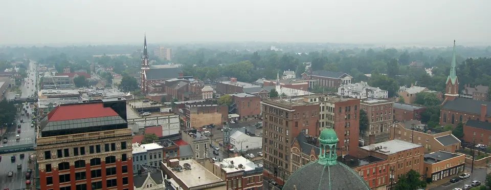 A wide rooftop view over downtown Lexington on a hazy, wet day in August 2006, with brick commercial buildings, a green copper church dome and two church steeples rising above the tree line.