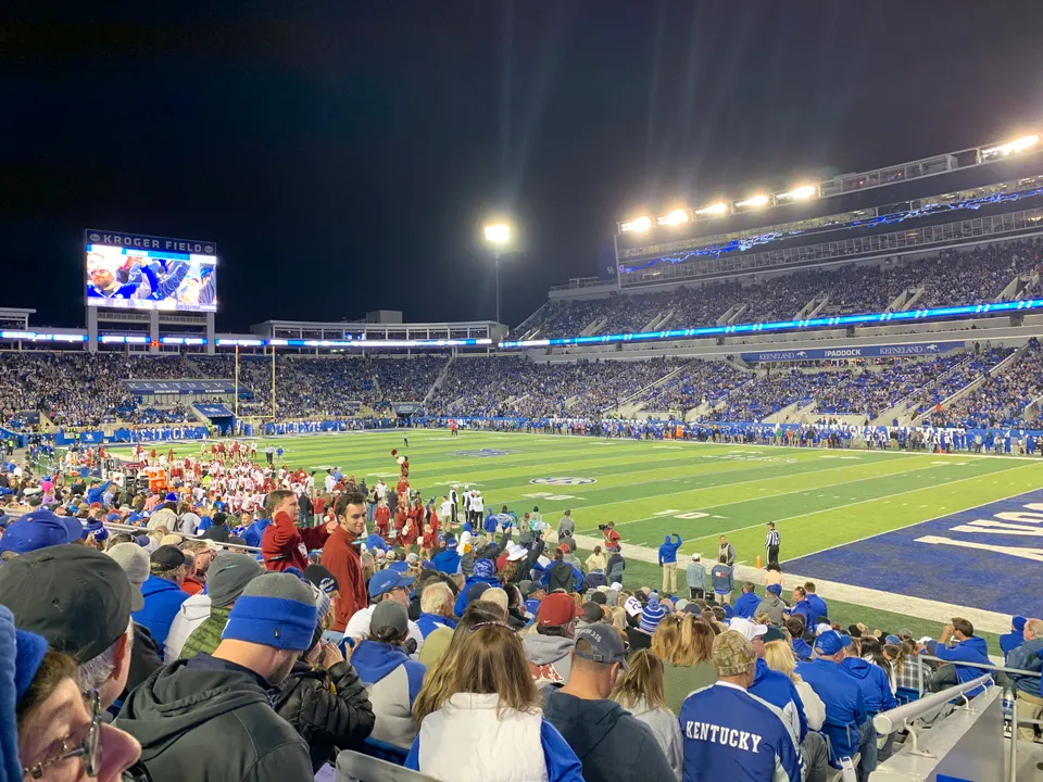 Kroger Field at night during the Kentucky vs. Arkansas football game in October 2019, seen from the lower stands: fans in blue in the foreground, the lit field and sidelines, the full east stands and the Kroger Field video board.