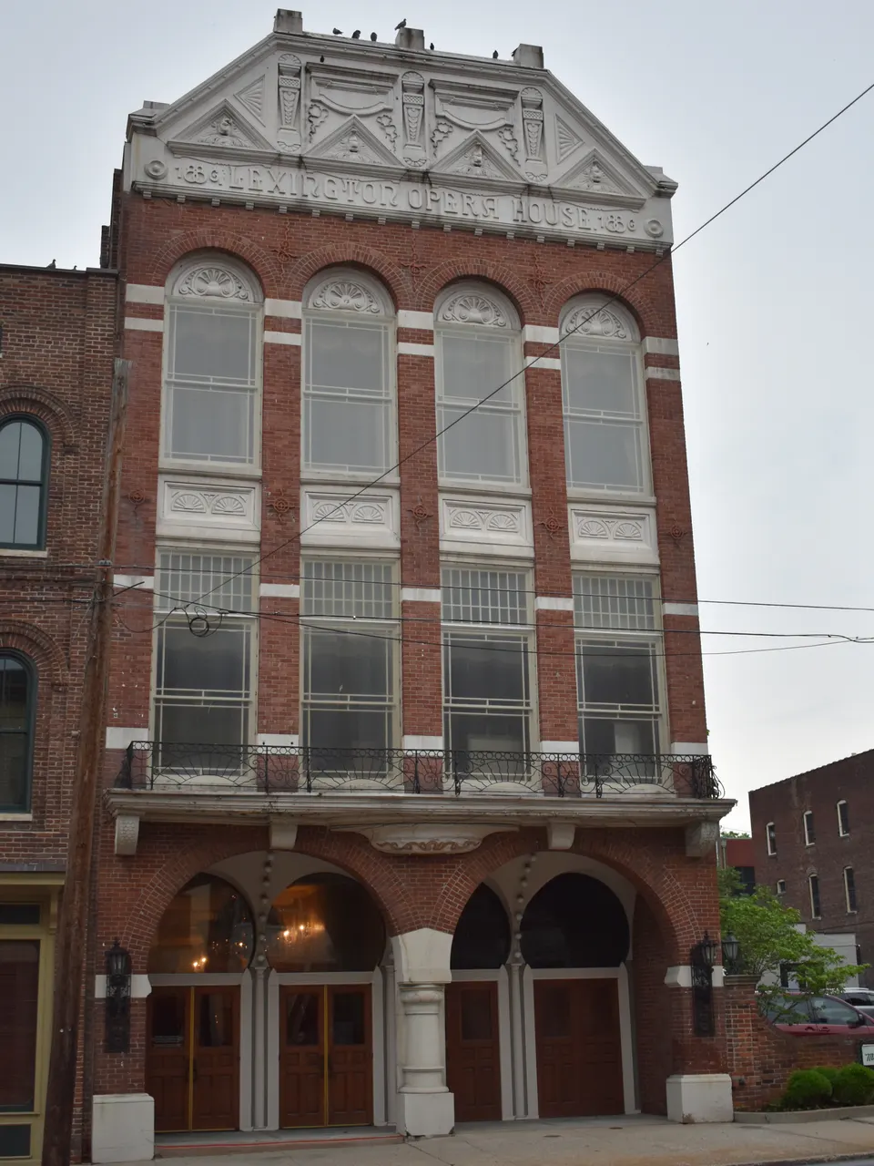 The red brick Lexington Opera House facade with its carved stone pediment, tall arched windows, iron balcony and arched entrance doors, seen from the street on an overcast day in June 2019.