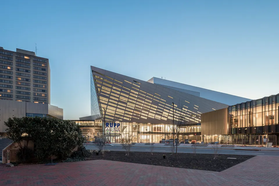 Rupp Arena's angled glass and metal facade lit from inside at dusk, seen across West High Street from the brick plaza at Triangle Park in January 2023, with bare young trees and fresh mulch in the foreground.