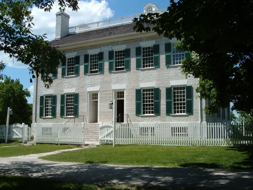 The limestone Centre Family Dwelling at Shaker Village of Pleasant Hill on a sunny day in May 2005, with green shutters, twin front doors, a white picket fence and a leafy tree in the foreground.